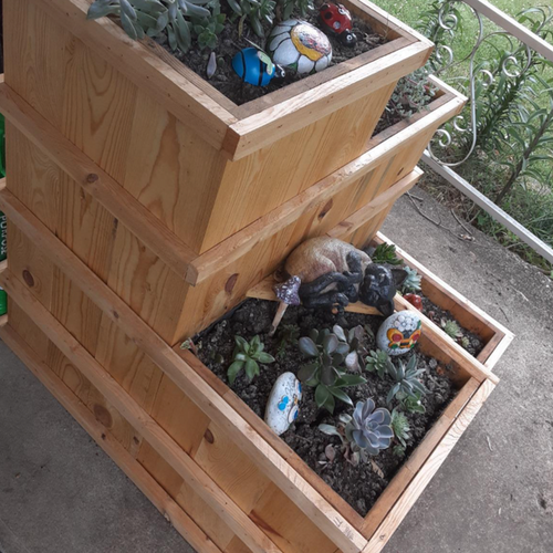 A stacked wooden planter filled with succulents and decorative painted stones, featuring a small cat sculpture nestled among the plants.