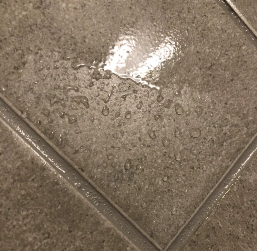Close-up of wet gray tile floor with visible water droplets and reflections, emphasizing the tiles' texture and sheen.