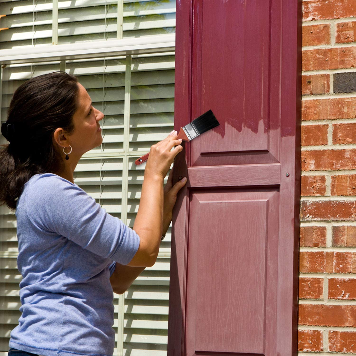 A person paints a wooden door with a paintbrush, standing beside a brick wall and window with white blinds.