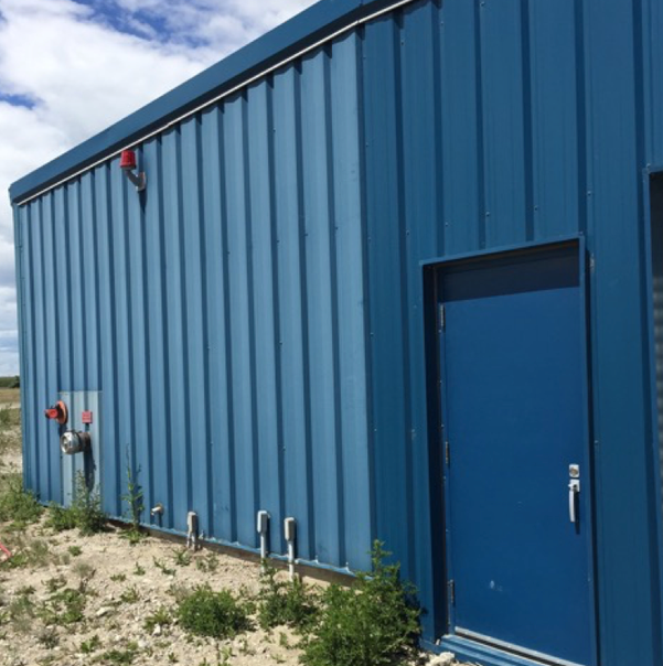 Side view of a blue metal building with a doorway, fire hydrant connection, and small weeds growing in the gravel below.