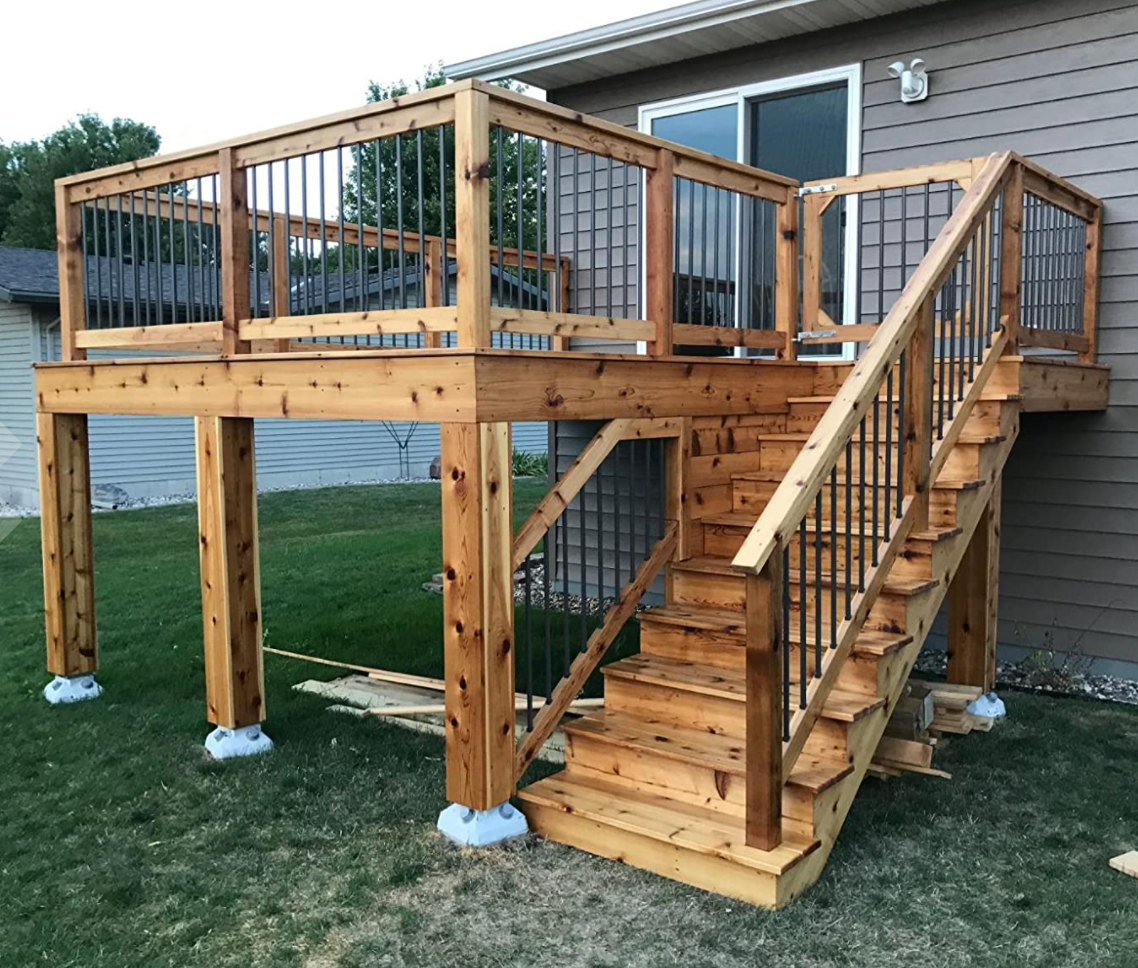 A wooden deck with a railing and stairs, elevated off the ground, beside a house with gray siding and a large window.