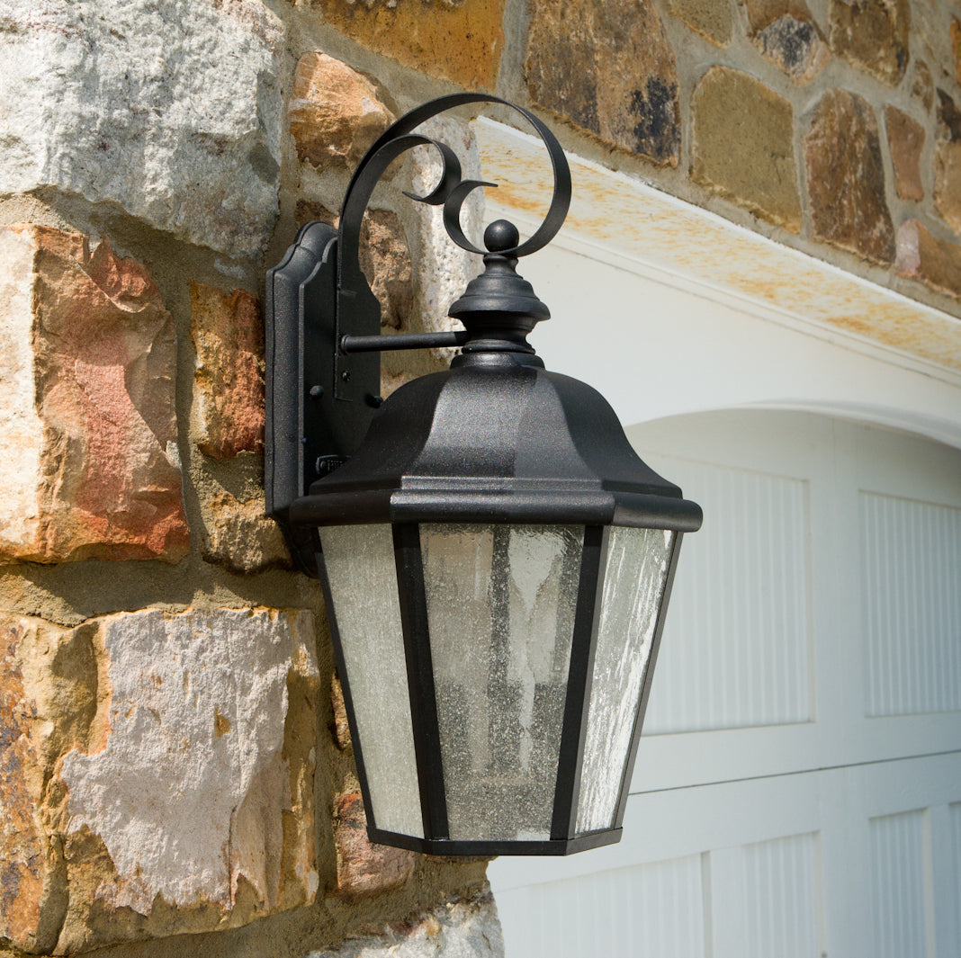 A decorative black lantern mounted on a stone wall, featuring a curved arm and glass panels, illuminating a charming entrance.