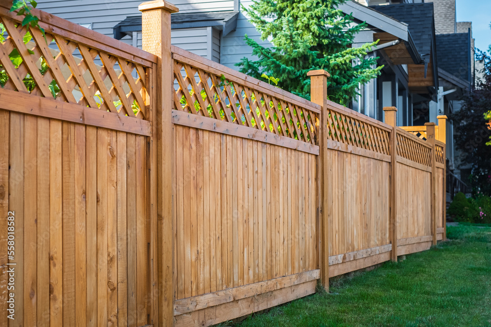 A long, wooden fence with a lattice top stretches along a grassy yard, bordered by trees and houses in a bright, sunny setting.