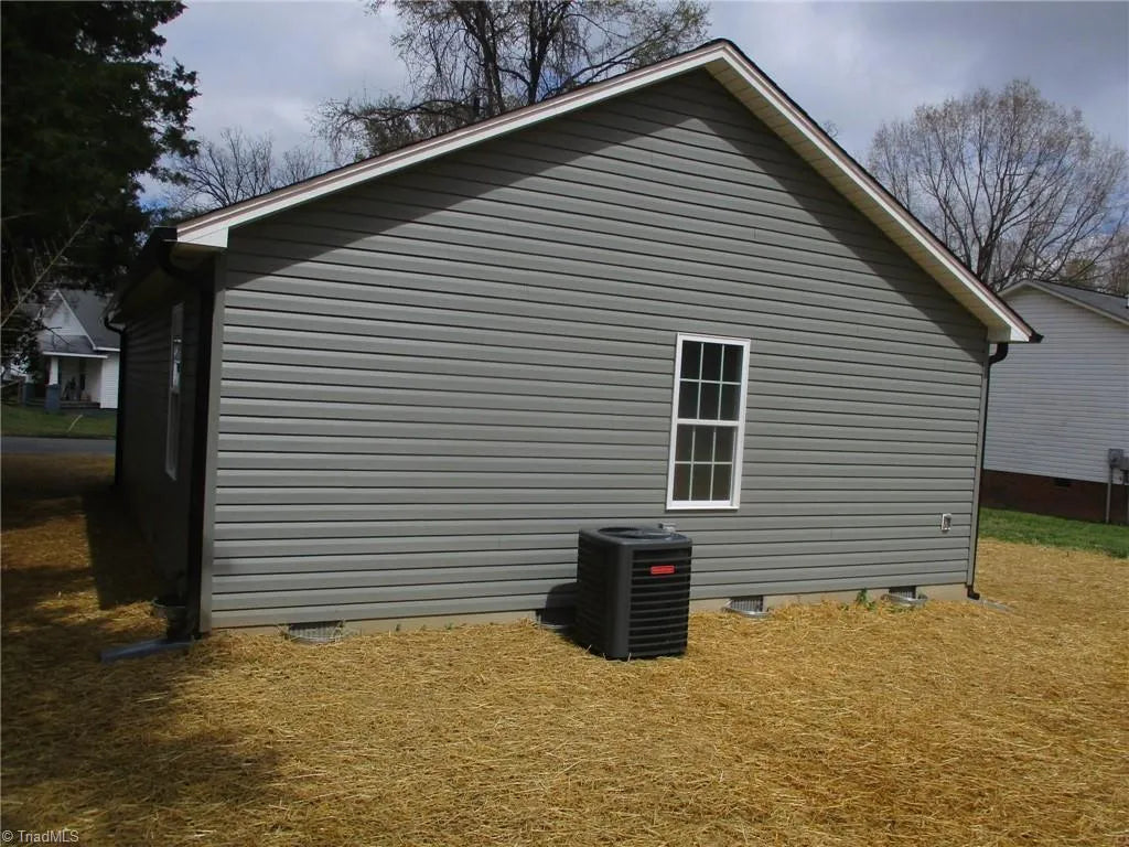 Side view of a gray vinyl-sided house with a small black air conditioning unit and straw covering the ground.