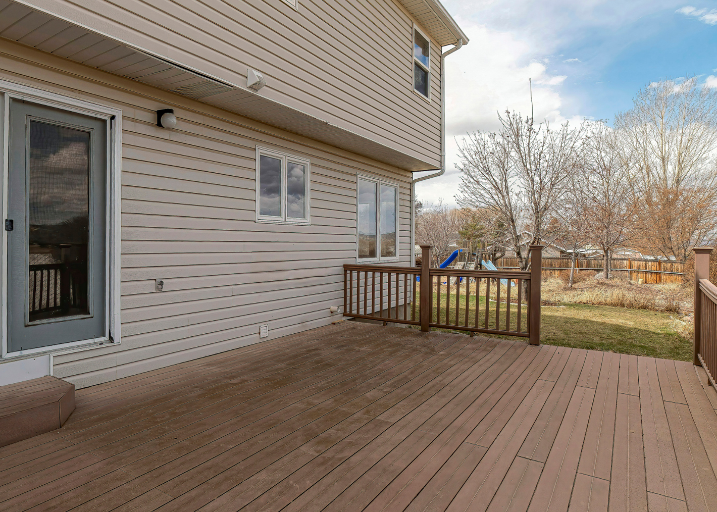 Wooden deck at the back of a house, featuring a door and railing, with a grassy yard and trees in the background.