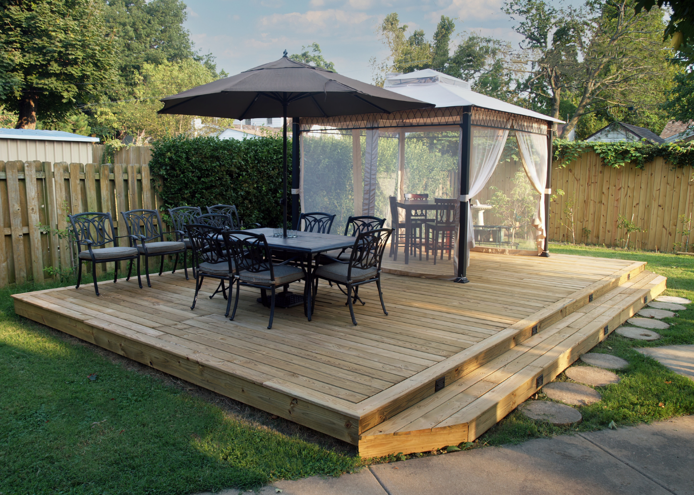 Wooden deck with a dining table and chairs, shaded by an umbrella, adjacent to a gazebo and surrounded by greenery.