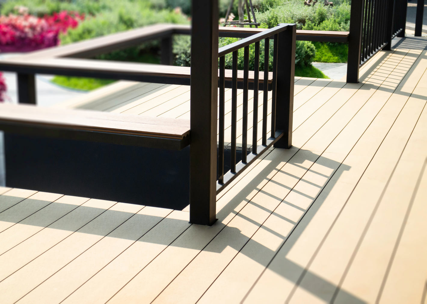 Close-up of a wooden deck with a black railing and shadows cast by the railing, surrounded by vibrant greenery and flowers in the background.