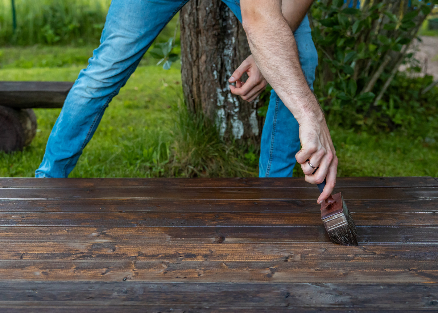 A person wearing blue jeans applies wood stain to a dark wooden table outdoors, with greenery in the background.